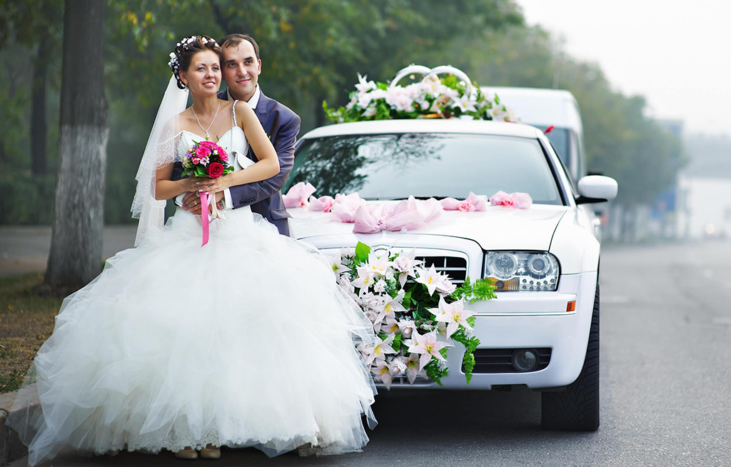 A bride in white dress with flowers and a groom in blue coat are smiling in front of a white limousine acquired from wedding limo rental service